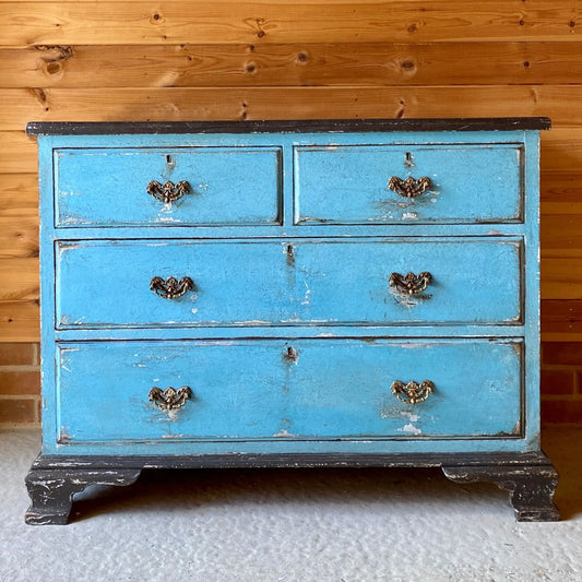 A worn blue dresser with three drawers and two smaller top drawers against a wooden wall. Features ornate handles and distressed, vintage charm.