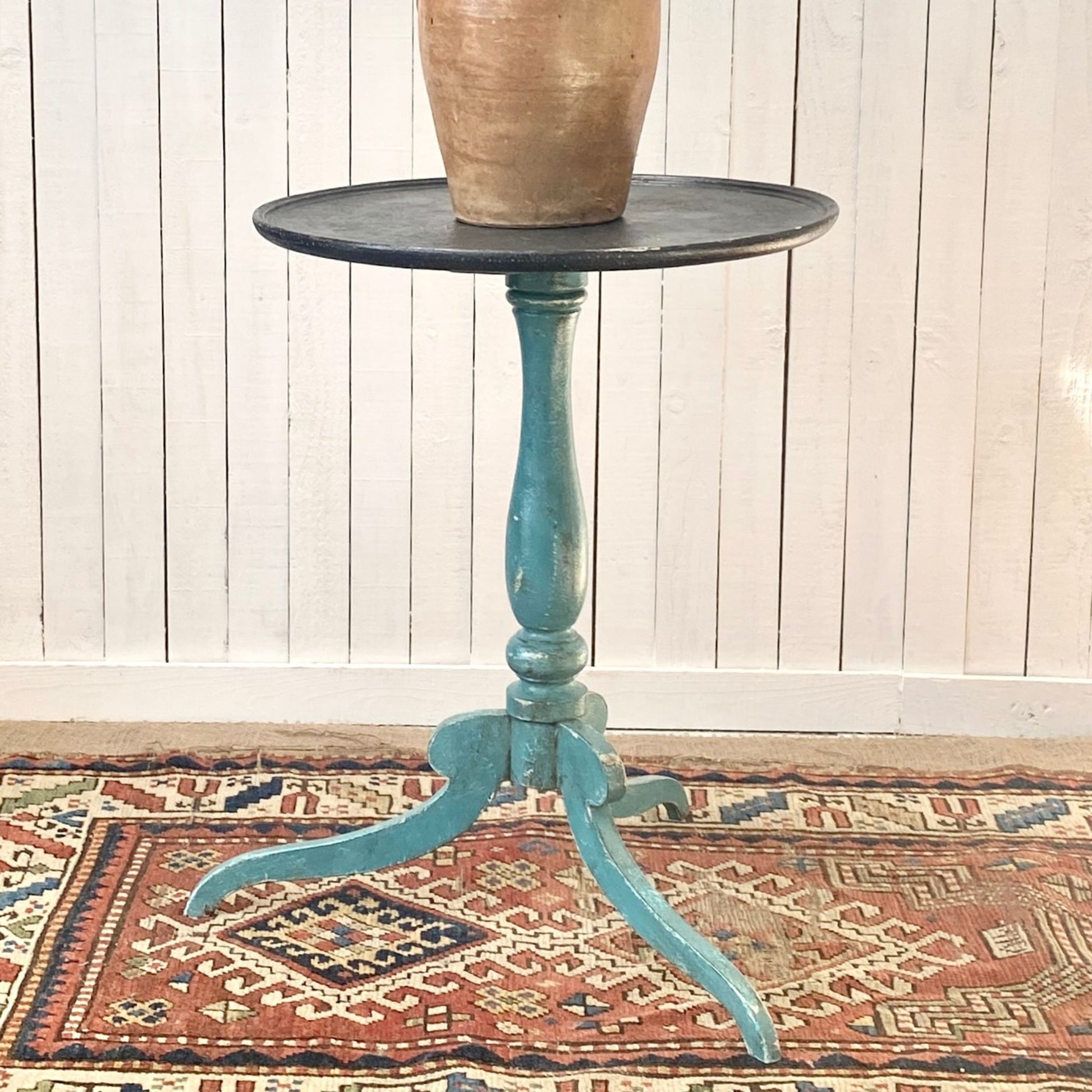 Small round pedestal wine table with a painted blue base and black top on a patterned rug against a wooden panel wall.